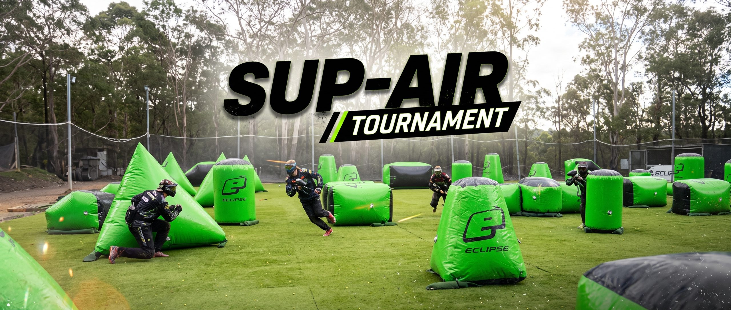 Action-packed panoramic photo of an outdoor tournament paintball field with bright green artificial turf and numerous inflatable green and black Sup-Air bunkers. Several players in full protective gear and jerseys are shown in motion—crouching, sprinting, and shooting—during an active game. High-contrast, sporty text in the upper-center reads "SUP-AIR / TOURNAMENT". A dense forest of eucalyptus trees and safety netting enclose the field under an overcast, natural sky.