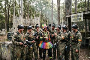 A group of men celebrating a bucks party at Action Paintball Yarramundi, laughing and gathered around the groom-to-be who is wearing a colorful tutu and costume over camouflaged paintball gear, with outdoor bushland fields and a sign reading "Action Paintball Yarramundi" in the background.
