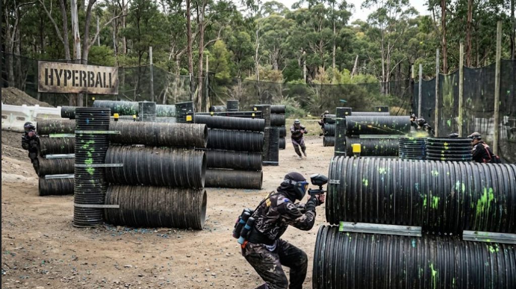 A paintball match in progress on the outdoor Hyperball field at Action Paintball Games Yarramundi. A player in dark camouflage and a blue-accented mask aims a marker from behind stacked black corrugated pipe bunkers splattered with bright green paint. Additional players navigate the dirt field in the background near safety netting, surrounding eucalyptus trees, and a large sign reading "HYPERBALL".